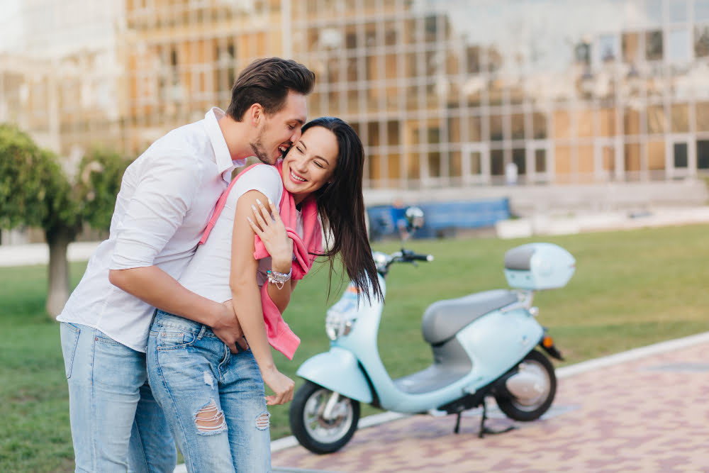 long haired smiling girl ripped blue jeans having fun with boyfriend city park outdoor portrait funny loving couple fooling around street with scooter background